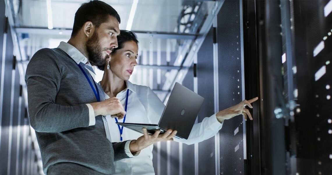 A man holding a laptop standing next to a woman pointing at a control panel
