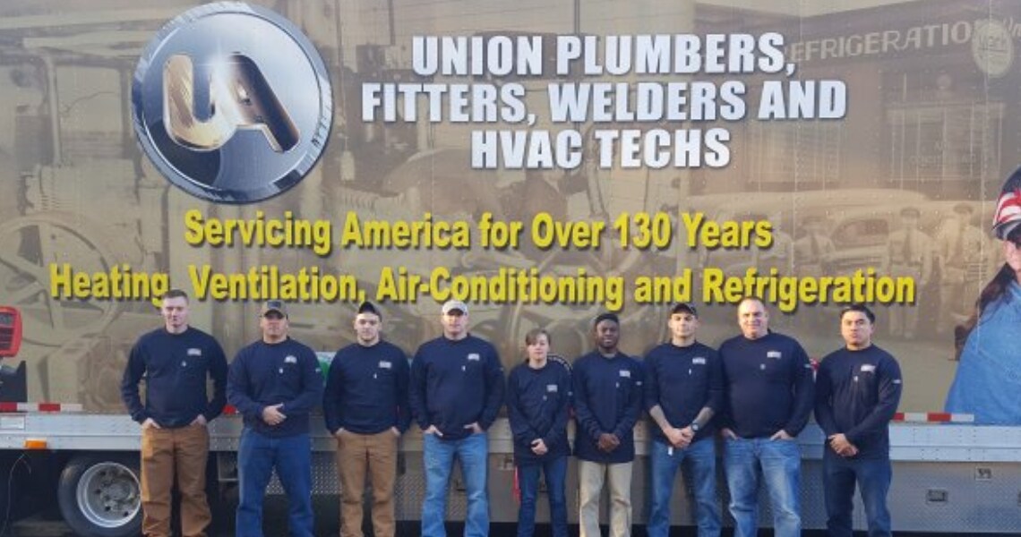 A group of plumbers posing in front of a sign that says 'Union Plumbers, Fitters, Welders and HVAC Techs'