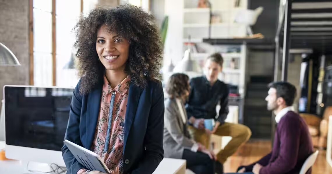 A female corporate employee holding a tablet and smiling, while her colleagues engage in a discussion in the background