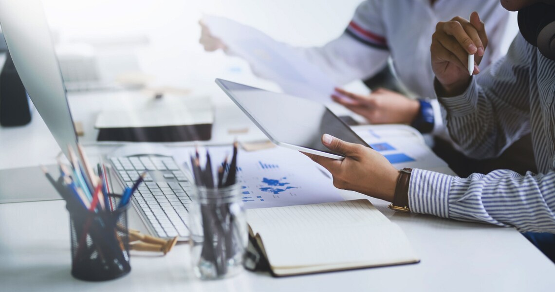 Close-up of people at a table with laptops and documents, while one of them holds a screen tablet