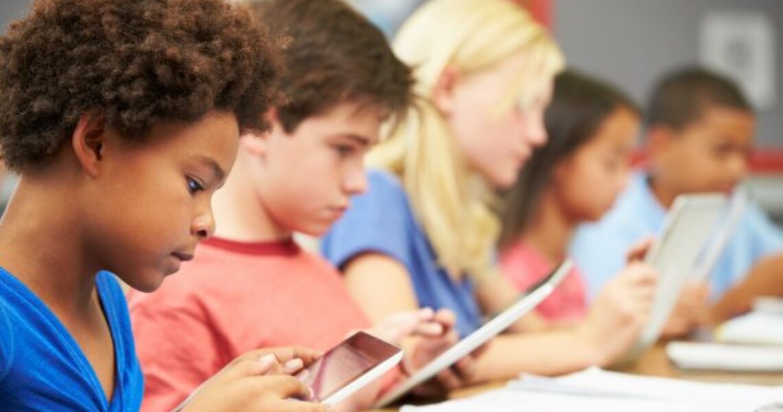 Young students sitting in a row at a table and looking at screen tablets