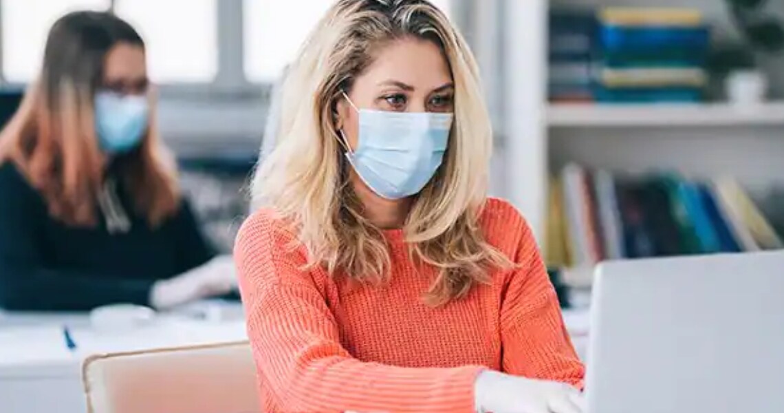 A woman wearing a facemask and scrolling on a laptop