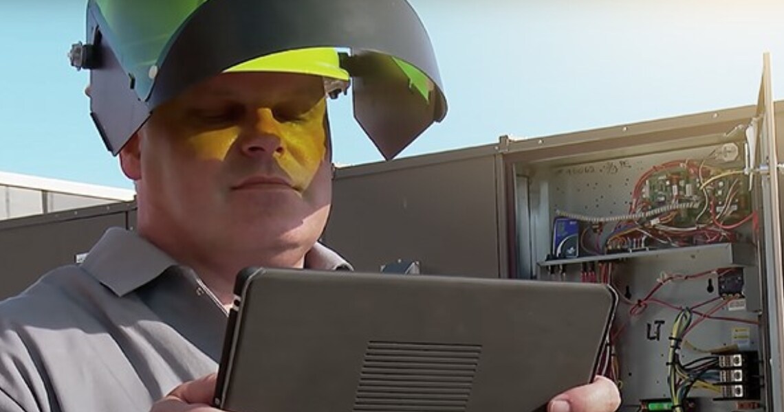 A technician wearing a helmet and looking at a tablet, in front of an air handling unit