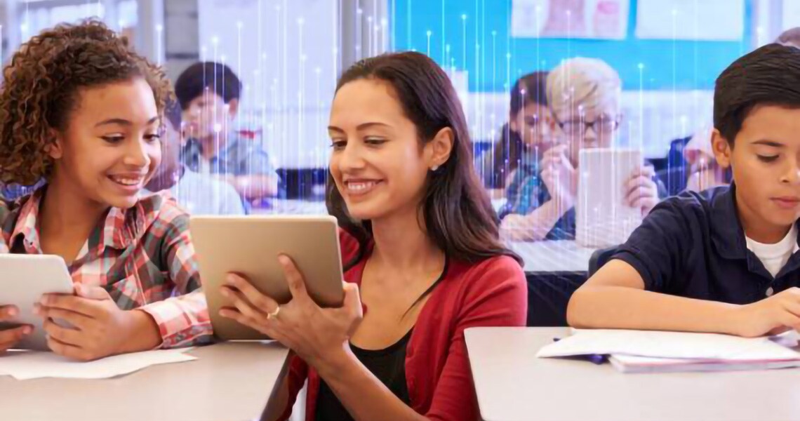 A teacher kneeling beside a young student and showing something on a tablet, in a classroom