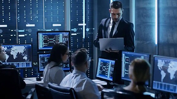 A group of people seated around desktops, with a man standing and holding a laptop, in an office