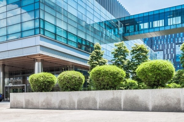 Large modern office building with blue-tinted glass walls, neatly maintained shrubs and trees, covered entrance area, and open paved space in front.