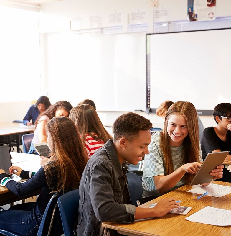 Two students with tablet laughing at each other while other students engaging on their work.