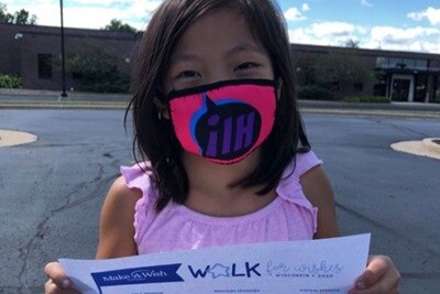 Grace, a young girl, wearing a facemask and holding up a 'Walk for Wishes' sign